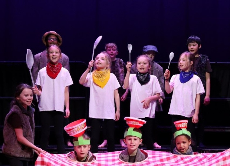 children with heads poking out of tablecloth and porridge bowls on their heads plus children in white t shirts and holding wooden spoons 